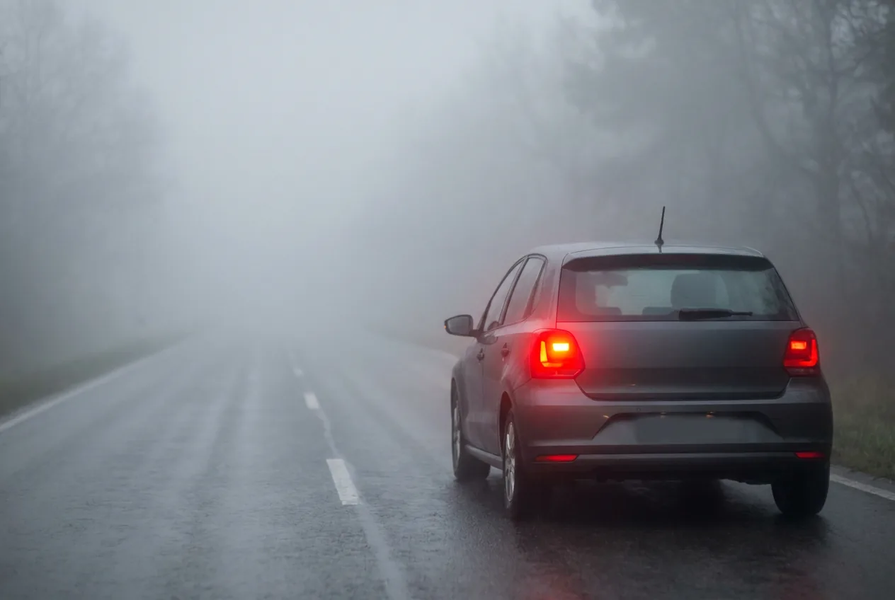 Coche visto desde atrás en niebla muy densa, con el entorno disuelto en gris y una luz roja intensa encendida en la parte trasera, más brillante que los pilotos que tiene al lado