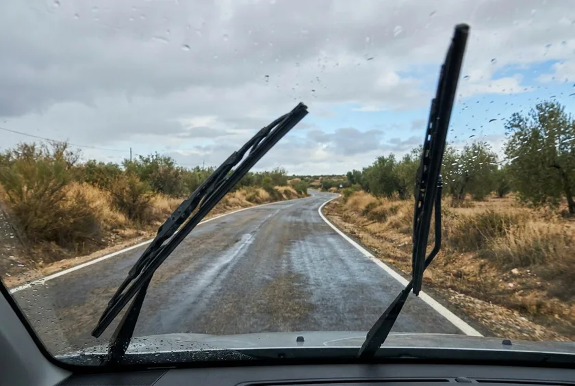 Vista desde el interior de un coche con los limpiaparabrisas en marcha sobre una carretera que empieza a mojarse con las primeras gotas de lluvia