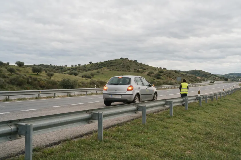 Persona con chaleco reflectante situada detrás del quitamiedos, junto a su vehículo averiado en el arcén