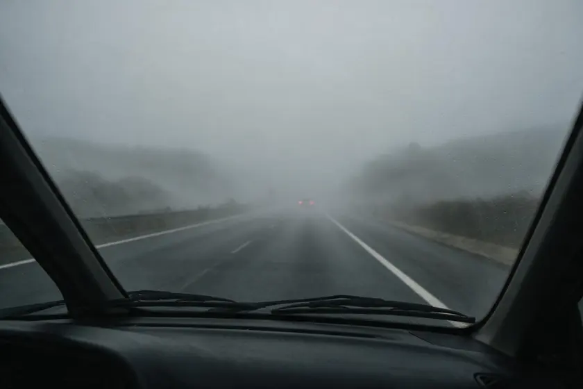 Carretera vista desde el puesto del conductor con niebla densa: la vía se pierde a pocos metros y solo se distinguen las luces traseras del vehículo de delante