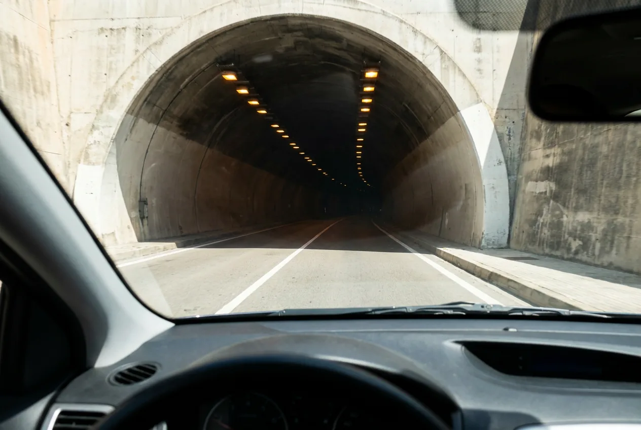 Vista desde el interior de un coche al entrar en un túnel de carretera, con las luces anaranjadas del techo alejándose y una acera elevada a la derecha