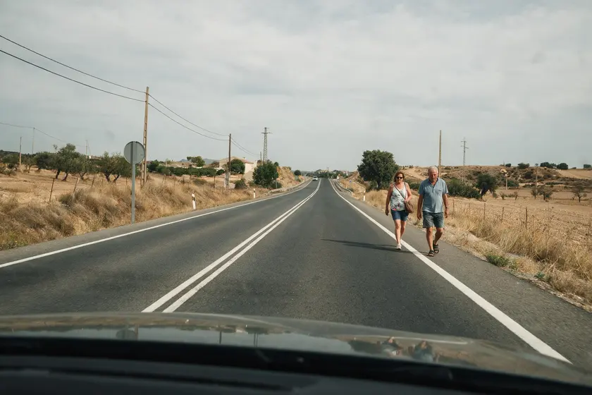 Dos peatones caminando por el lado izquierdo de una carretera, de frente al tráfico que se acerca