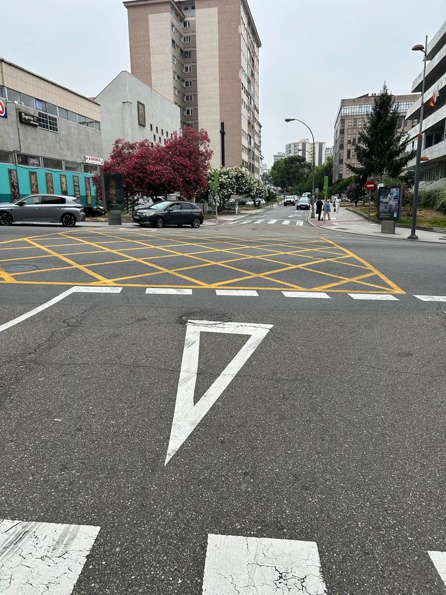 Cruce urbano con una cuadrícula de líneas amarillas entrecruzadas pintada sobre la calzada y un triángulo blanco pintado en el carril, con el vértice hacia el conductor