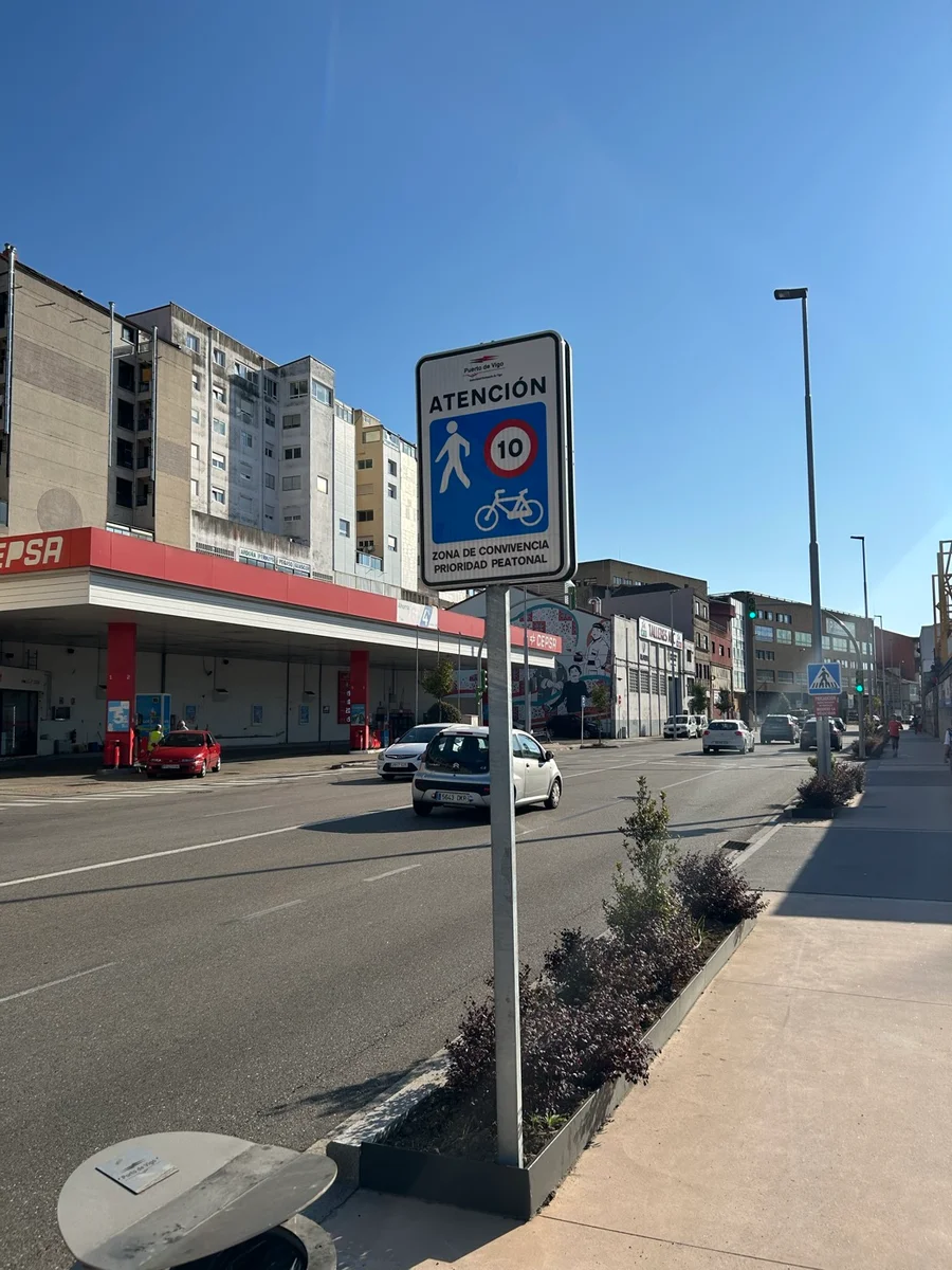 Señal cuadrada azul con una escena de calle: casas, personas caminando y un coche